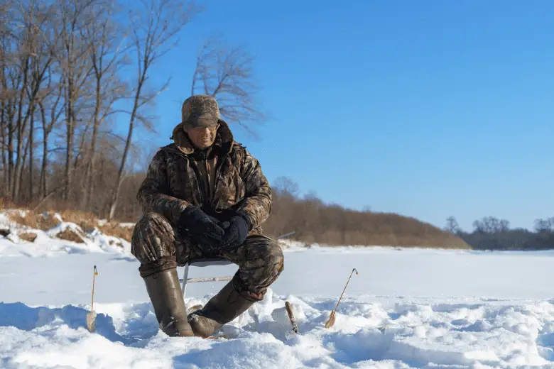 A man is fishing close to Ice hole at winter sunny day under blue sky